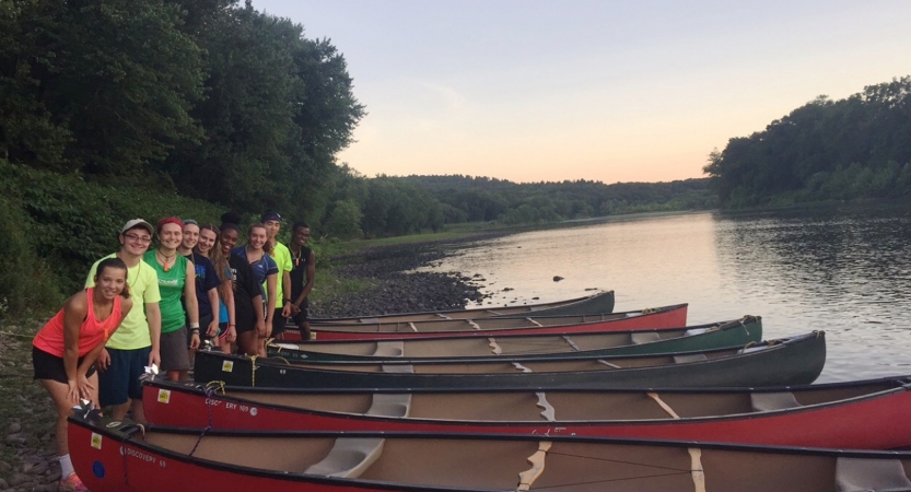 A group of people stand beside beached canoes beside a calm river